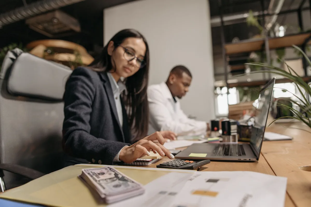 Two colleagues working at a shared desk with a laptop, a calculator and financial papers in an office