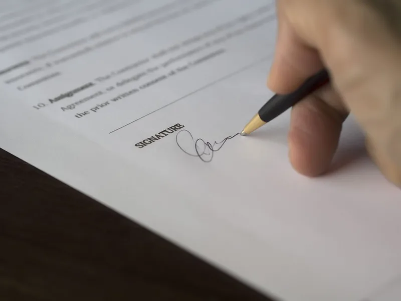 A hand signing a printed agreement above the signature line with a black pen