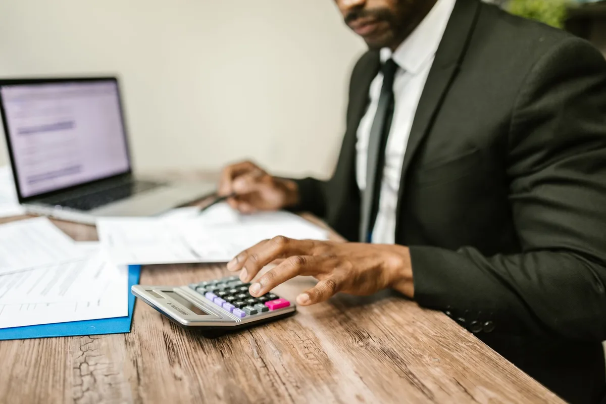 A person in a dark suit using a calculator beside a laptop and printed financial documents on a wooden desk