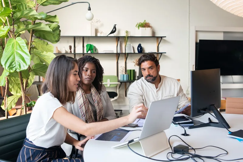 Three colleagues gathered around a laptop in a bright office, discussing figures on screen