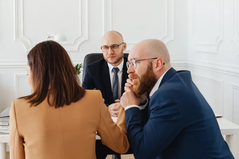Two men and a woman in business clothes talking across a white desk in a bright office