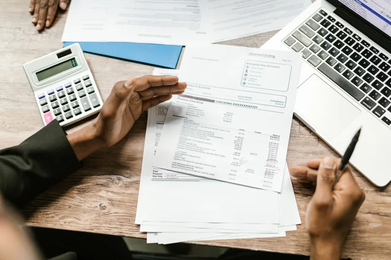 Hands holding a bank statement beside a calculator, a laptop and a pen on a wooden desk